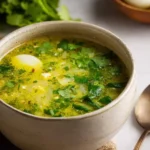A rustic bowl of vibrant Green Borscht, garnished with fresh herbs and a soft-boiled egg, on a textured surface.
