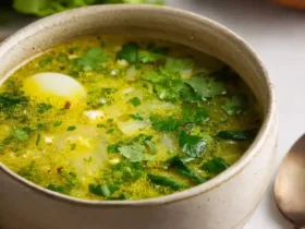 A rustic bowl of vibrant Green Borscht, garnished with fresh herbs and a soft-boiled egg, on a textured surface.