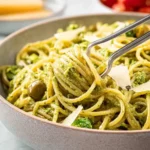 Close-up of spaghetti tossed in a vibrant green olive pasta sauce, garnished with parmesan and olives, being served with metal tongs.