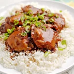 Glistening Hawaiian Shoyu Chicken thighs on a bed of white rice, garnished with fresh green onions and sesame seeds, on a white plate.