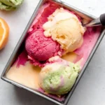 Scoops of vibrant Homemade Rainbow Sherbet in pink, orange, and green flavors fill a metal pan, with an ice cream scoop.
