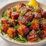 A close-up shot of a delicious Honey Garlic Salmon Bites Bowl, garnished with sesame seeds, green onions, and a lemon wedge.