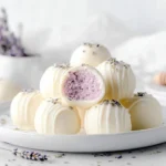 A pile of Honey Lavender White Chocolate Truffles on a white plate, with one truffle cut in half to reveal its purple filling, garnished with lavender buds.
