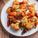 A close-up shot of a white plate filled with golden-fried Kung Pao Cauliflower, garnished with red chilies and green onions.