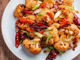 A close-up shot of a white plate filled with golden-fried Kung Pao Cauliflower, garnished with red chilies and green onions.