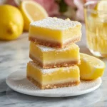 A stack of three golden lemon bars topped with powdered sugar, alongside fresh lemons and a glass of lemonade.