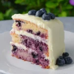 A delightful slice of Lemon Blueberry Cake with Cream Cheese Frosting on a white plate, showcasing moist cake layers, vibrant blueberries, and rich white frosting.