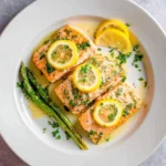 Three flaky pieces of lemon butter salmon garnished with fresh parsley and lemon slices, served with roasted asparagus on a white plate.