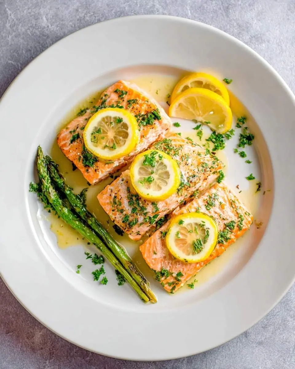 lemon butter salmon 29 Three flaky pieces of lemon butter salmon garnished with fresh parsley and lemon slices, served with roasted asparagus on a white plate.