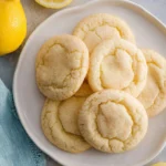 A white plate filled with light-colored, crinkled lemon cookies, dusted with a light spice, surrounded by fresh whole and sliced lemons and a blue napkin.