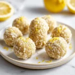 A plate of delicious Lemon Energy Balls coated in shredded coconut and topped with fresh lemon zest, with whole and halved lemons in the background.