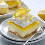 A close-up of a vibrant slice of lemon lush dessert on a white plate, topped with fluffy whipped cream, lemon zest, and a lemon slice.