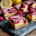 Close-up of a delicious Lemon Raspberry Bar with a vibrant swirled topping on black slate, with more bars and fresh lemons in the background.