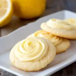 A white plate holds several delicious Lemon Sugar Cookies topped with swirled lemon frosting, with fresh lemons blurred in the background.