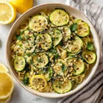 A vibrant bowl of Lemon Zucchini Pasta garnished with fresh zucchini slices, toasted pine nuts, herbs, and chili flakes, alongside fresh lemons.