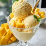 Three scoops of creamy mango and coconut ice cream in a glass sundae dish, garnished with fresh mango cubes and mint leaves, with a spoon.