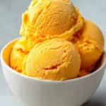A white bowl brimming with several scoops of vibrant mango ice cream on a light grey background.