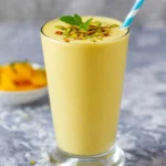 A vibrant Mango Lassi Smoothie in a tall glass, garnished with chopped pistachios, mint, and a blue and white straw, on a textured grey surface.
