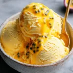 A close-up shot of creamy Mango Passionfruit Ice Cream scoops topped with a vibrant passionfruit sauce and seeds in a speckled bowl.