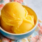 A blue bowl holds several scoops of bright yellow mango sorbet on a colorful floral napkin.