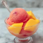 A refreshing glass bowl filled with scoops of vibrant red Mango Strawberry Sorbet and bright yellow mango chunks, with a spoon.