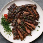 A platter of perfectly grilled Marinated Skirt Steak, thinly sliced and topped with fresh herbs, alongside cherry tomatoes.