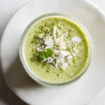 A vibrant green Matcha Coconut Smoothie served in a glass, garnished with shredded coconut, matcha powder, and a fresh mint sprig on a white plate.