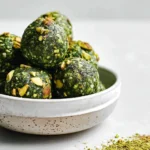 A white speckled bowl filled with vibrant green Matcha Pistachio Energy Balls, topped with chopped pistachios, with more green powder scattered on the side.