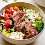 A vibrant mediterranean steak bowl featuring perfectly sliced medium-rare steak, fresh cucumber, tomato, red onion salad, feta, and tzatziki sauce.