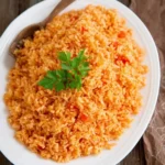 A white platter overflowing with vibrant orange Mexican Rice, garnished with fresh parsley and a wooden spoon, set on a rustic wooden table.