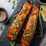 Two glistening miso glazed salmon fillets with sesame seeds and green onions on a black slate board, with lemons and sauce.