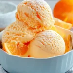 Three scoops of creamy Orange Creamsicle Ice Cream in a blue bowl, garnished with fresh orange slices and a dusting of powdered sugar.