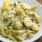 A white bowl filled with creamy Pasta with Artichokes and Lemon, garnished with fresh parsley, grated cheese, and a lemon wedge.