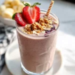 A refreshing Peanut Butter & Jelly Keto Smoothie in a clear glass, garnished with fresh strawberries, granola, and a striped straw.