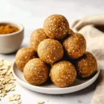 A pyramid stack of delicious Pumpkin Spice Protein Balls on a white plate with scattered oats and a bowl of spices in the background.