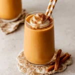 A delicious Pumpkin Spice Smoothie in a glass, garnished with whipped cream, cinnamon, and striped straws, next to cinnamon sticks.