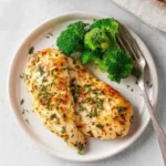A plate featuring two golden-brown Ranch Baked Chicken Breasts, seasoned with green herbs, served alongside vibrant steamed broccoli florets and a silver fork.