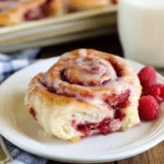A glazed raspberry cinnamon roll on a white plate, with a bite revealing the sweet fruit filling, fresh raspberries, and milk in the background.