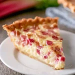 A vibrant slice of rhubarb custard pie with creamy filling and red rhubarb pieces sits on a white plate.
