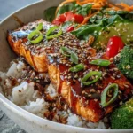 A close-up view of a delicious Salmon Teriyaki Bowl with perfectly glazed salmon, sesame seeds, green onions, white rice, and a vibrant side salad.