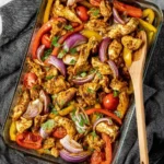 Overhead shot of a delicious Sheet Pan Chicken Shawarma with colorful bell peppers, red onions, and tomatoes in a glass baking dish with a wooden spoon.