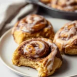 A close-up shows a partially eaten sourdough discard cinnamon roll with sweet white icing on a plate, surrounded by more rolls.