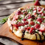 A delicious Strawberry Balsamic & Goat Cheese Pizza with fresh strawberries, crumbled goat cheese, basil, and balsamic glaze on a wooden board outdoors.