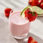 A vibrant pink Strawberry Coconut Smoothie in a clear glass, garnished with a fresh strawberry and lime slice on a light table.
