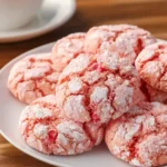 A close-up shot of a white plate piled high with delicious pink Strawberry Crinkle Cookies, dusted with powdered sugar, next to a blurred teacup.