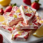 A vibrant pile of triangular Strawberry Lemon Frozen Yogurt Bark pieces, studded with strawberries and lemon zest, resting on a white marble slab with fresh fruit in the background.