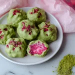 A plate of vibrant green Strawberry Matcha Clusters, one bitten to reveal a bright pink, textured freeze-dried strawberry interior, with matcha powder on the side.