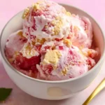 A white bowl holds several scoops of vibrant Strawberry Shortcake Ice Cream, swirled with red fruit and white cream, topped with golden shortbread crumbs.