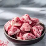A dark bowl brimming with delicious Sugar Free Frozen Yogurt Bites, dusted with vibrant red sprinkles, on a light marble surface.