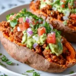 Close-up of two delicious Taco Stuffed Sweet Potatoes topped with guacamole, pico de gallo, and fresh cilantro on a white plate.